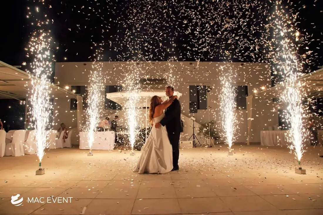 A bride and groom share their first dance on an outdoor terrace, surrounded by sparklers shooting into the night sky and falling confetti, creating a magical wedding moment.