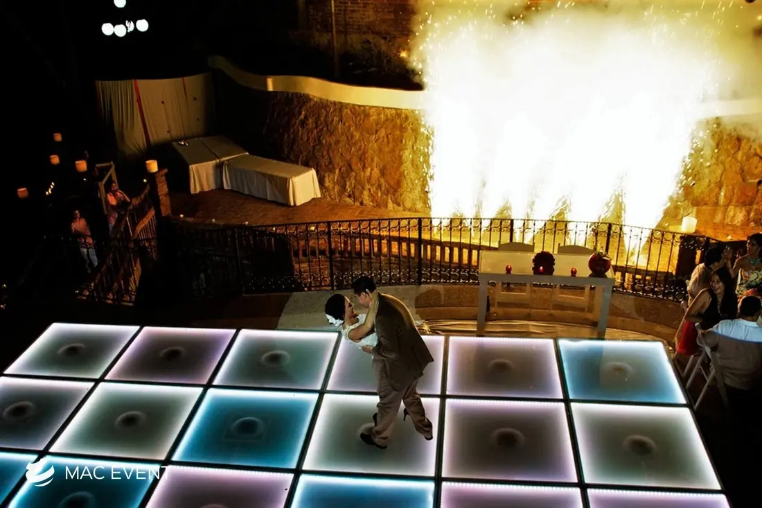 A couple performs a romantic dance on a glowing LED dance floor as white fireworks erupt behind them, illuminating the wedding venue with a golden glow.