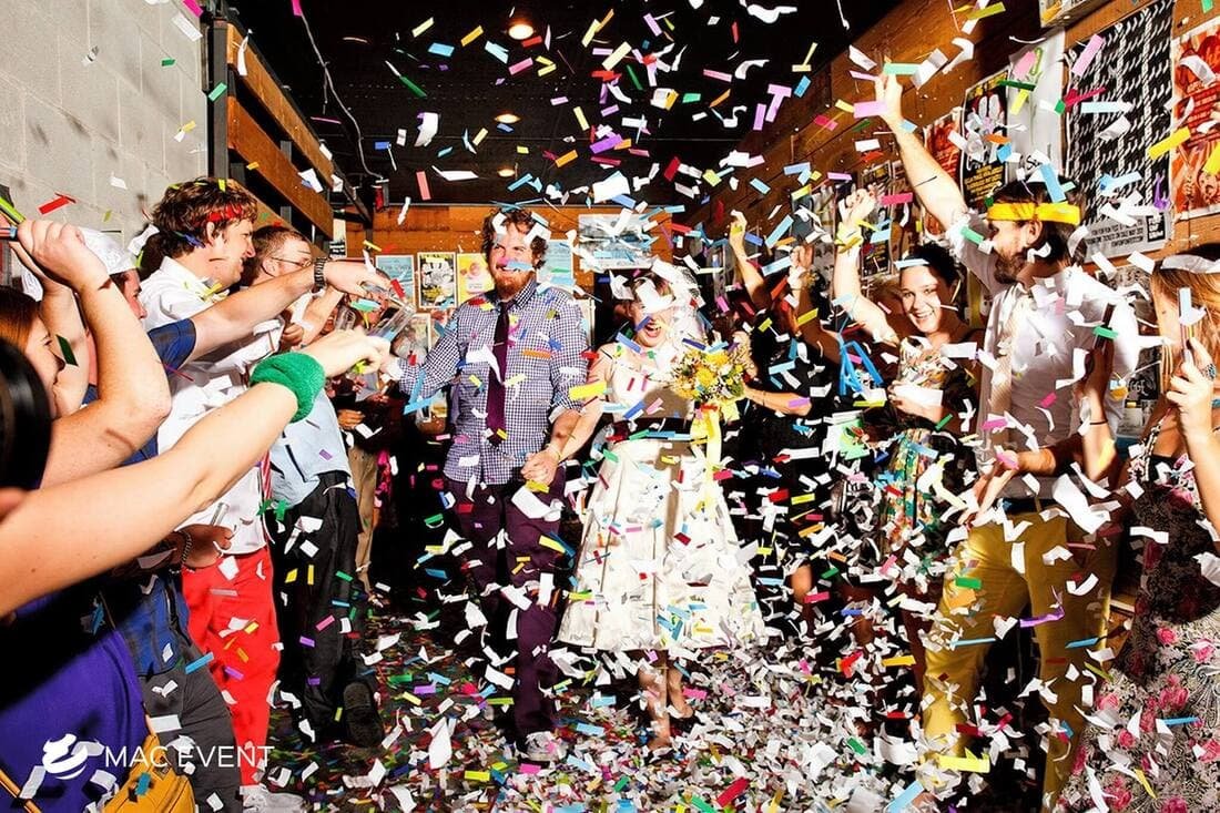 A newlywed couple walks through a lively crowd at their wedding reception, as friends and family shower them with multicolored confetti in a fun and festive atmosphere.